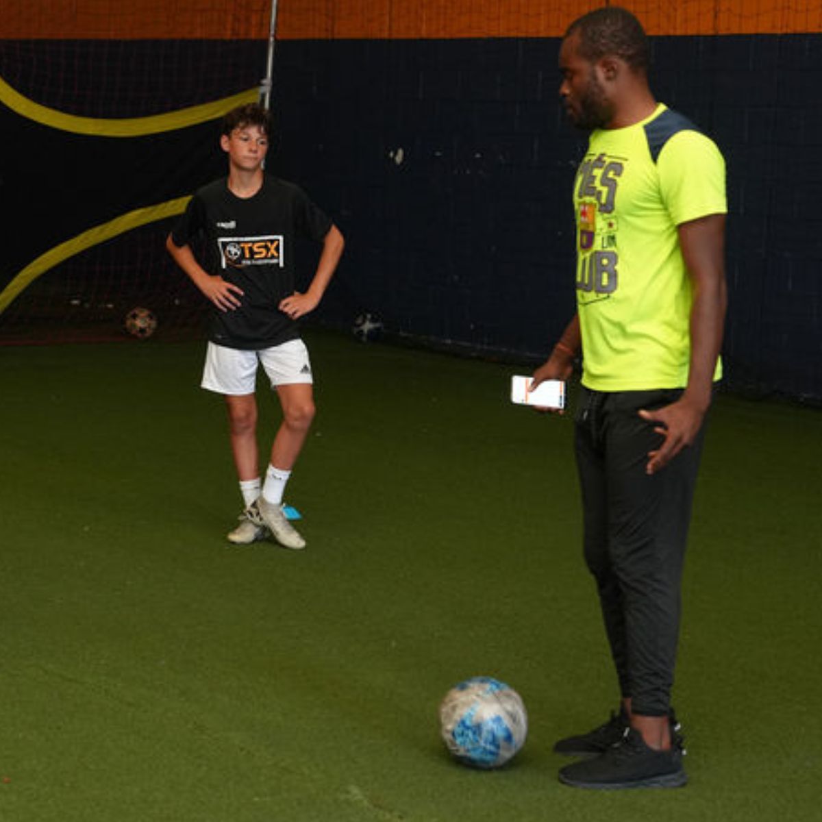 Total Soccer trainer and kid on a sports field with a soccer ball, one wearing a TSX shirt and the other in a bright yellow shirt.