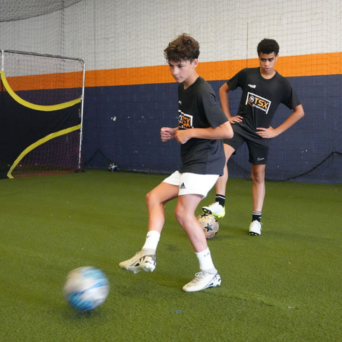 Total Soccer kids  playing soccer indoors on a green artificial turf with a blue and orange wall in the background.