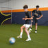 Total Soccer kids  playing soccer indoors on a green artificial turf with a blue and orange wall in the background.